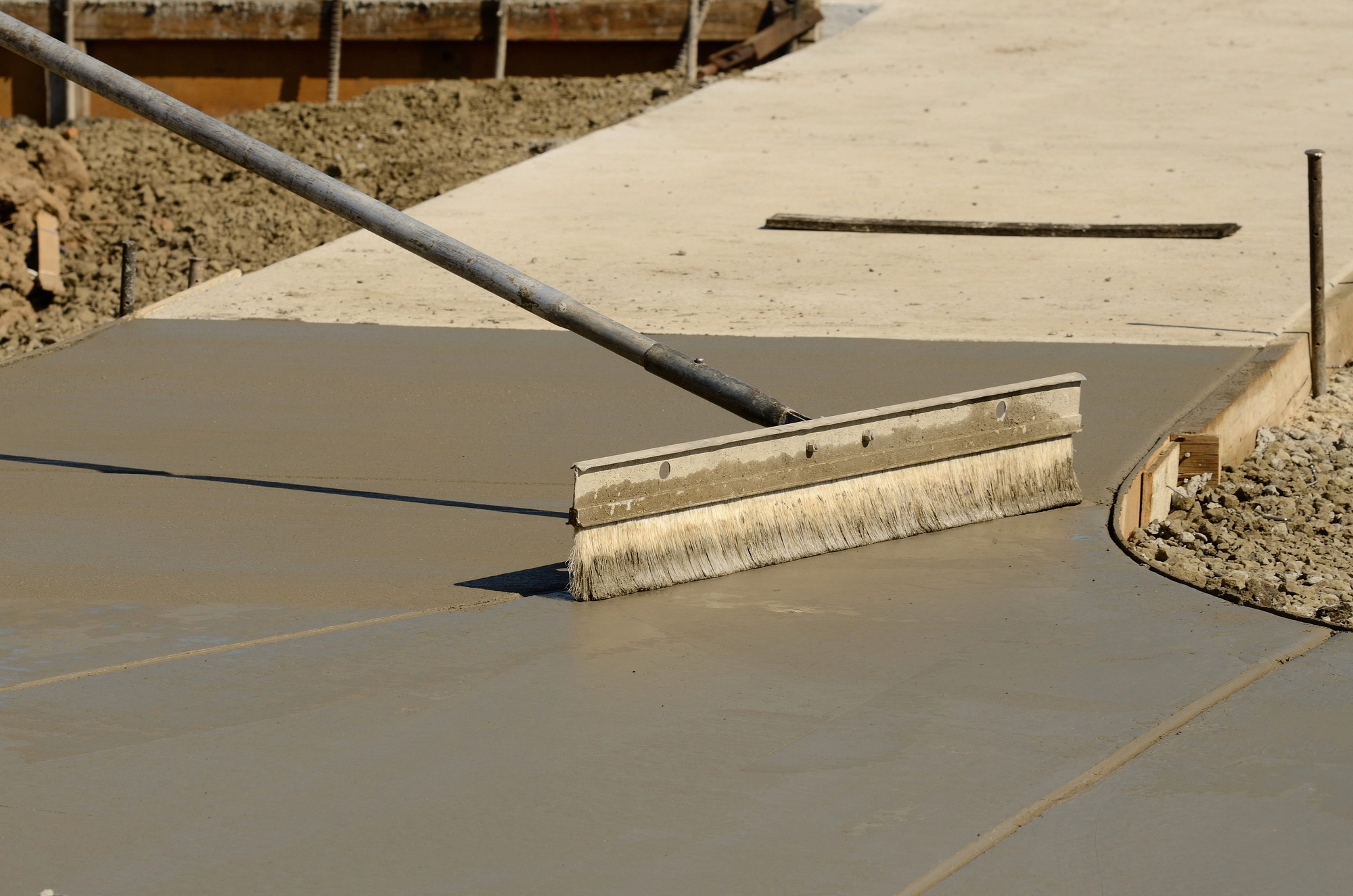 A large broom smooths and textures freshly poured concrete on a curved section of a sidewalk during construction.