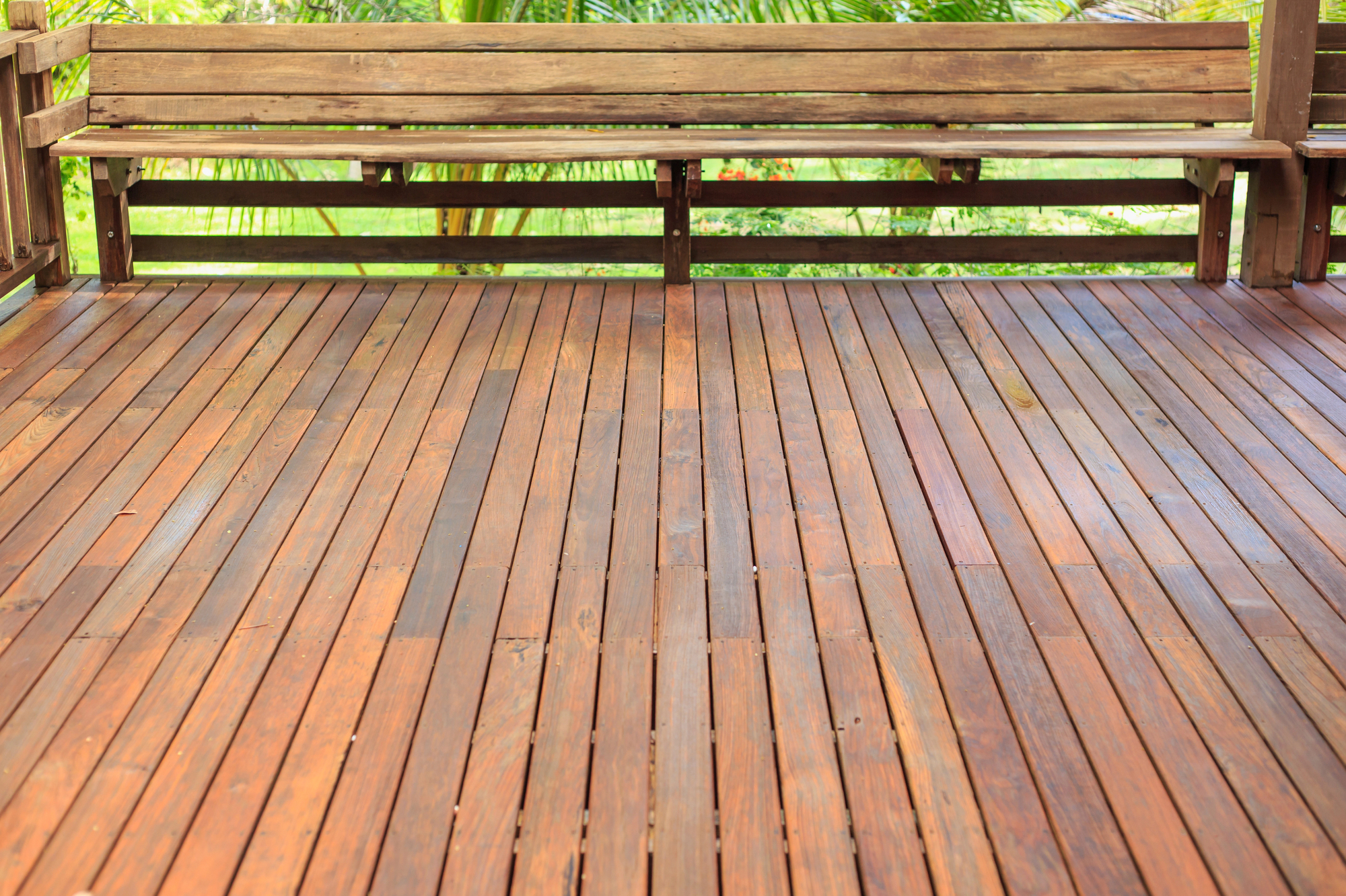 Wooden deck with built-in bench, photographed outdoors in daylight. Green foliage is visible beyond the deck.