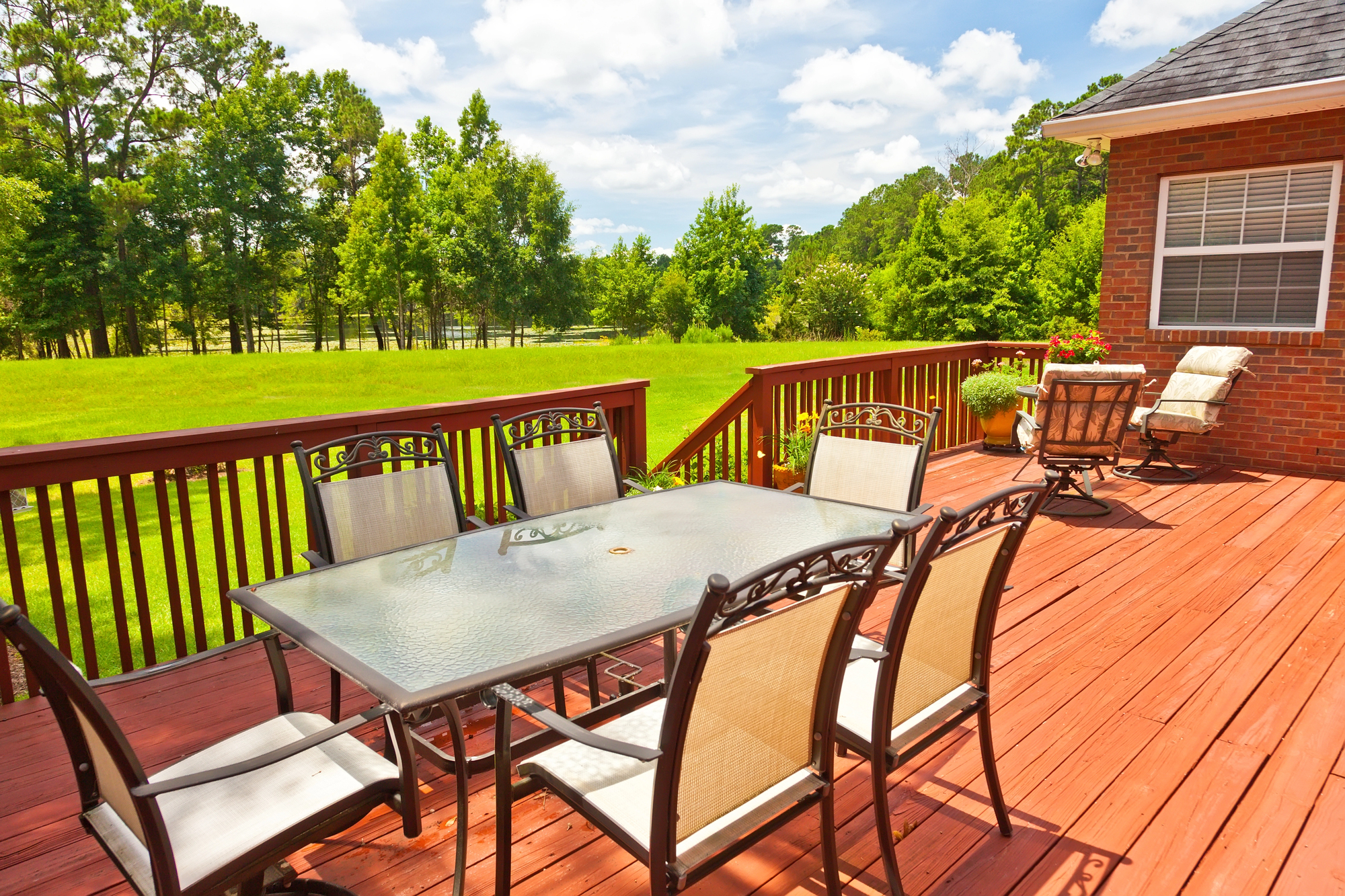 A glass-top table with six chairs sits on a wooden deck overlooking a grassy backyard with trees and a partly cloudy sky.