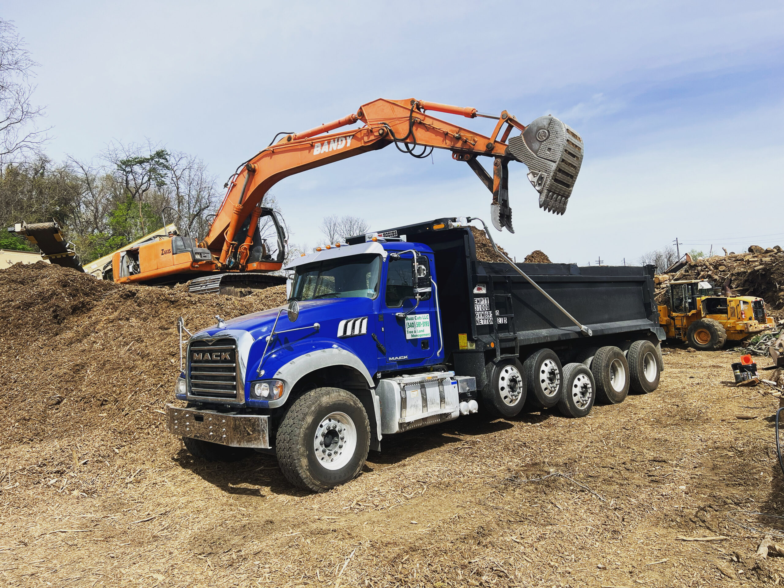 An excavator loads dirt into a blue dump truck at a construction site with piles of soil and other heavy equipment in the background.