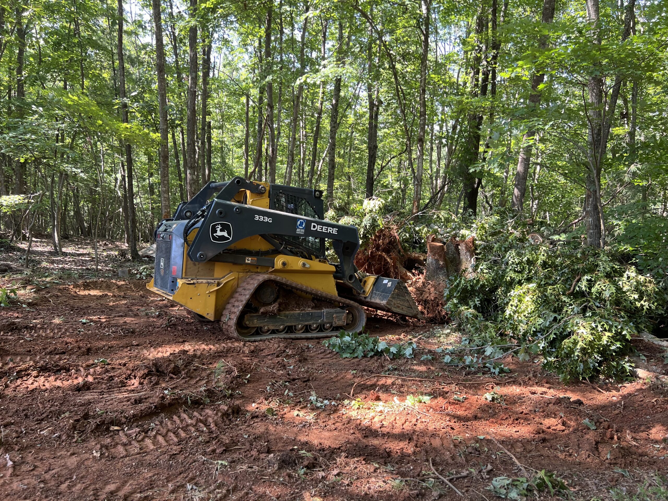 A John Deere compact track loader clears fallen trees and debris in a forested area, leaving tracks in the reddish soil.