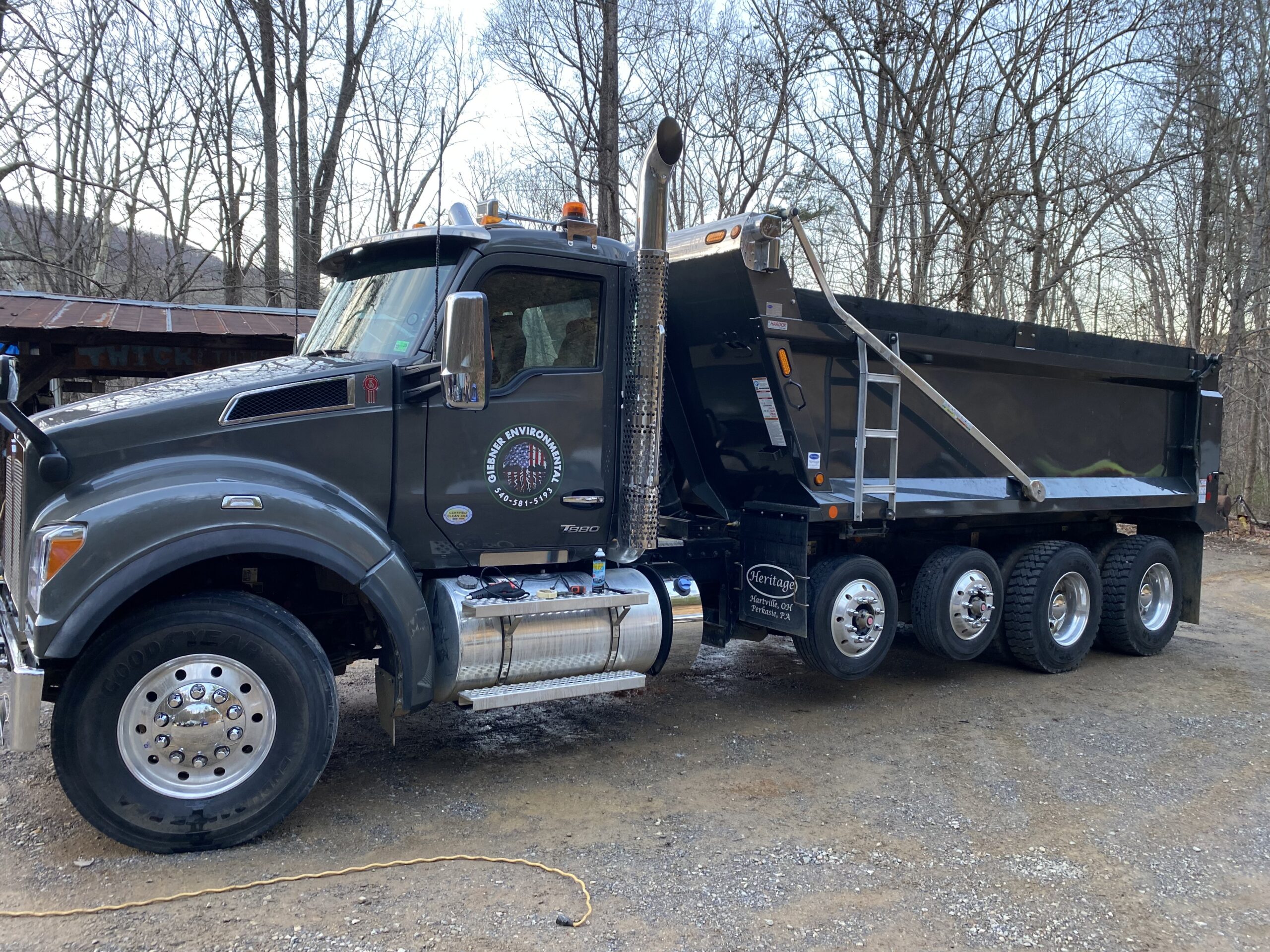 Black dump truck with four rear axles is parked on a gravel lot surrounded by bare trees in a wooded area.