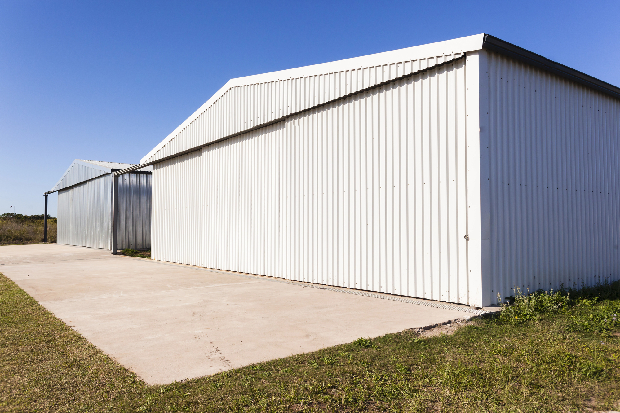 Two large white metal storage buildings with sliding doors stand side by side on a concrete pad under a clear blue sky. Grass and dirt border the concrete at the front and side.