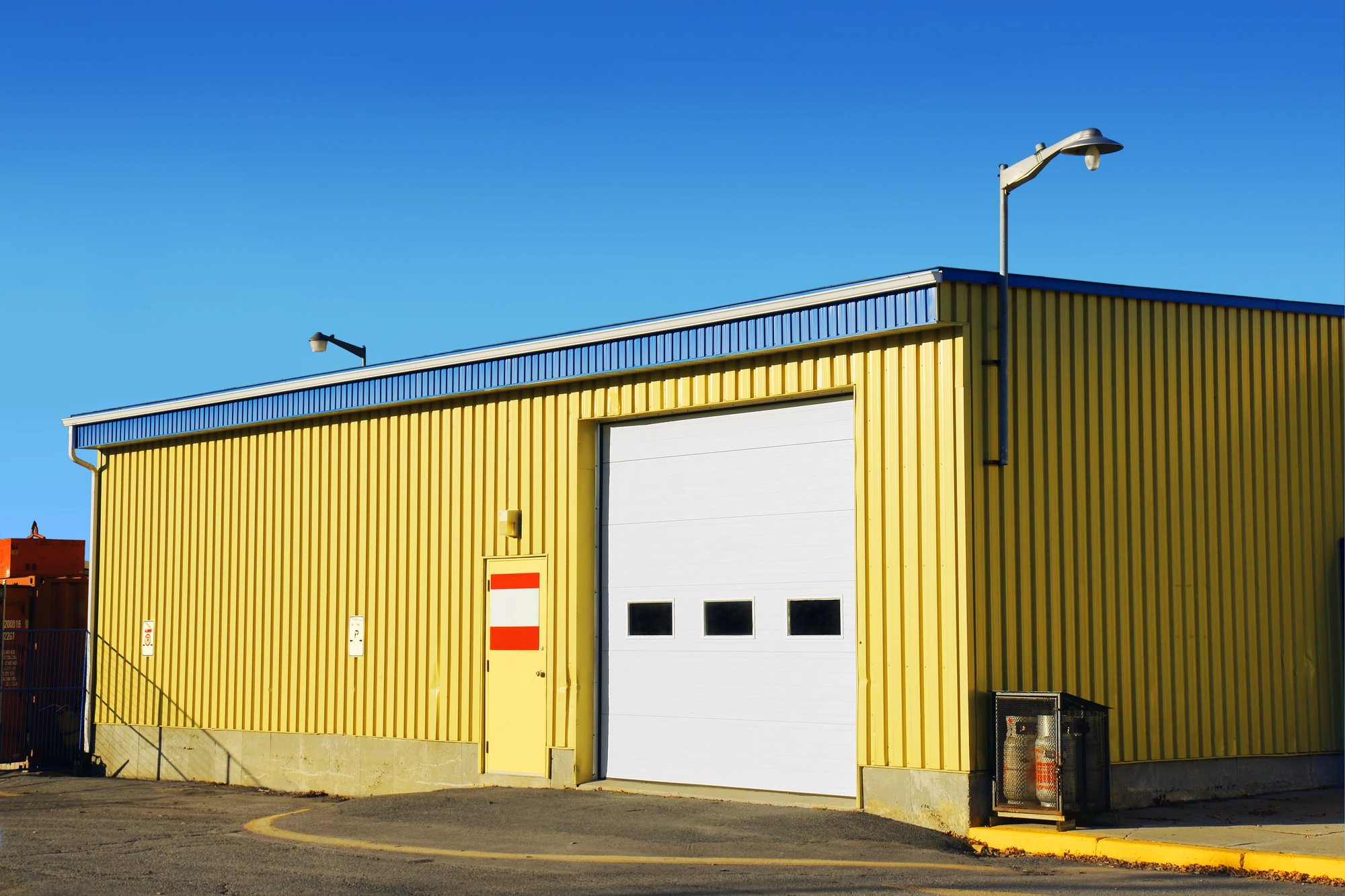 A yellow industrial building with a large white garage door, red and white signs, outdoor lights, and a clear blue sky overhead.