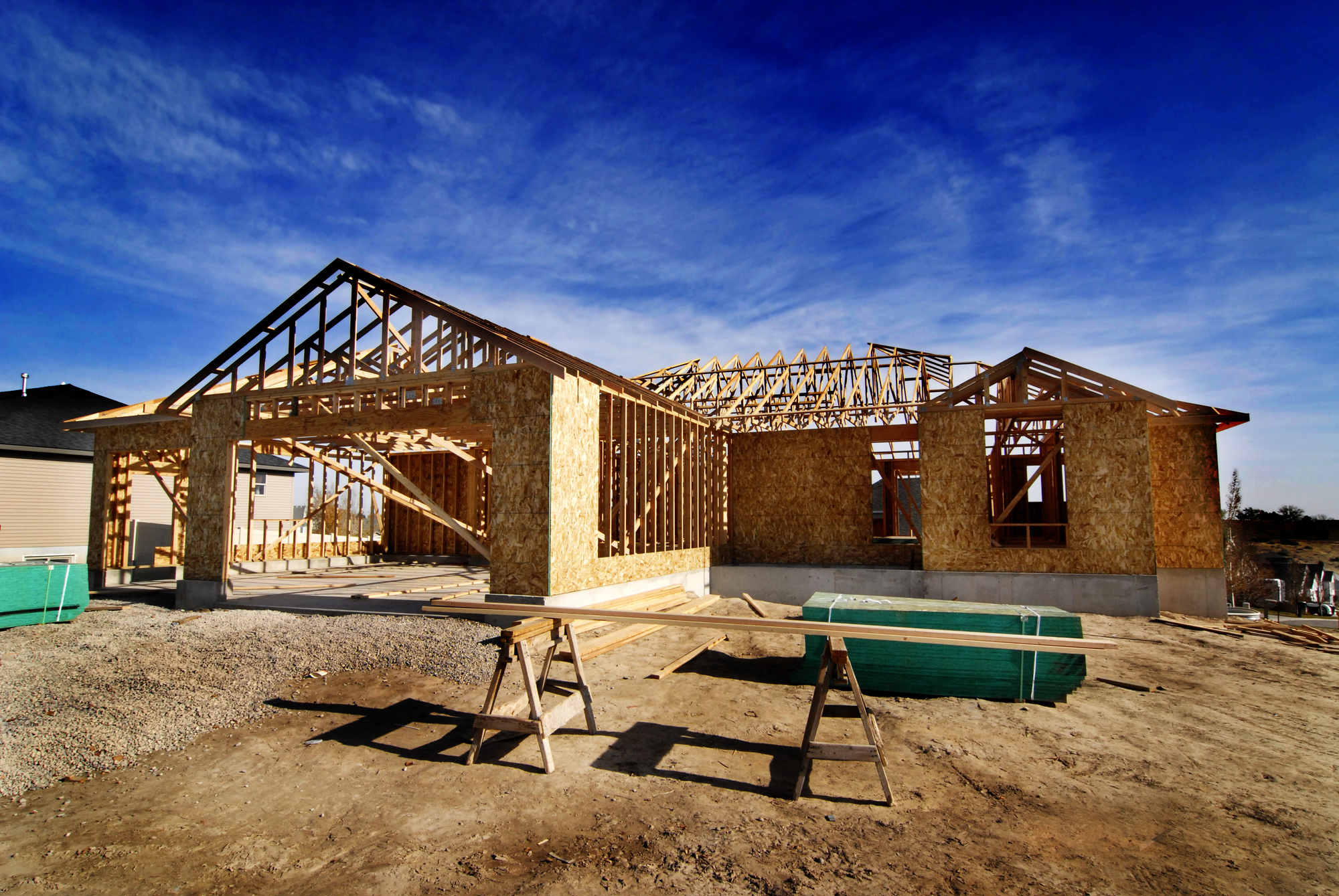 Partially constructed house with exposed wooden framework and plywood walls under a blue sky, surrounded by construction materials and dirt.