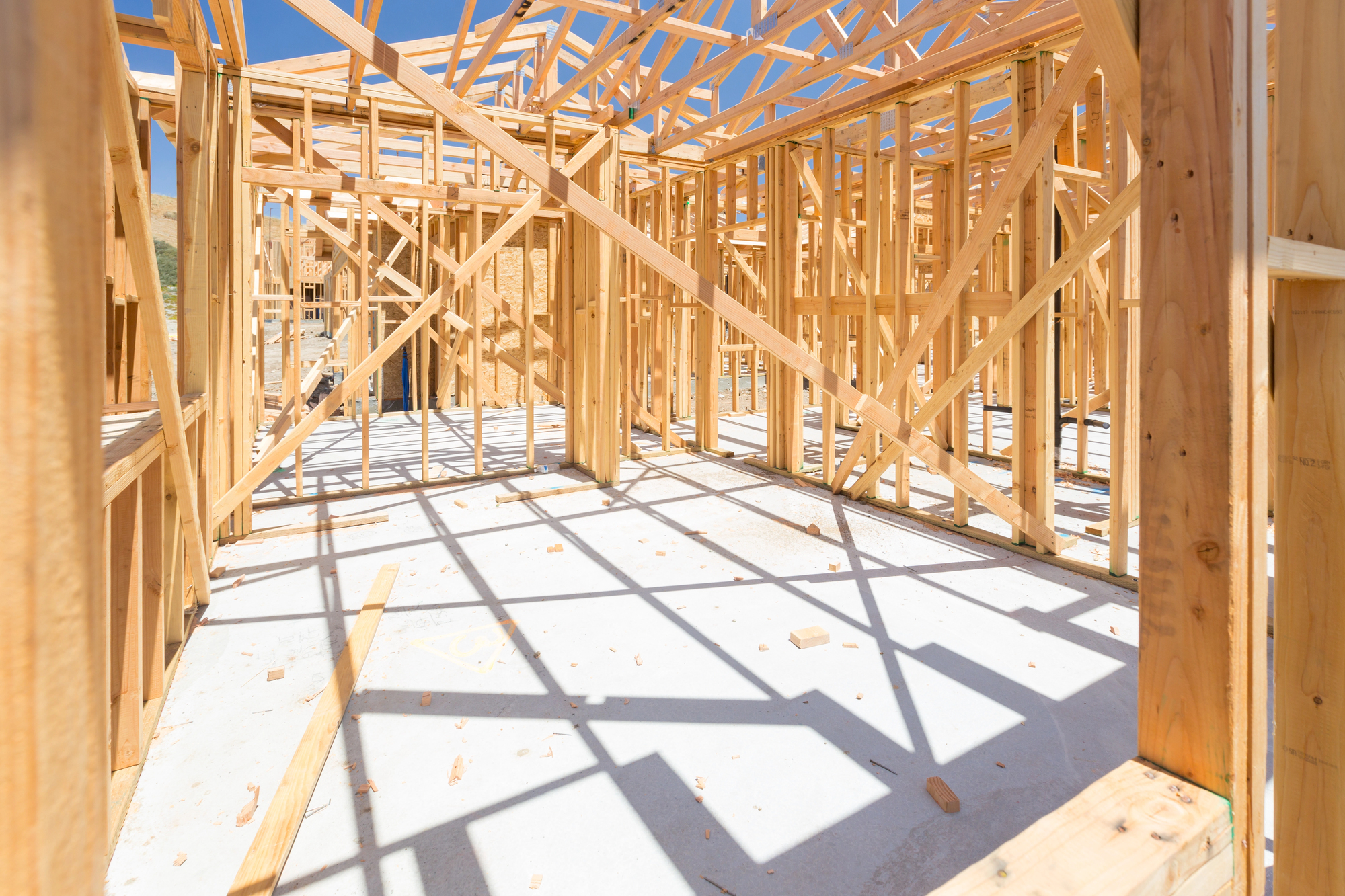 Wooden framing for a house under construction, with exposed beams and supports on a concrete foundation, seen in bright sunlight.