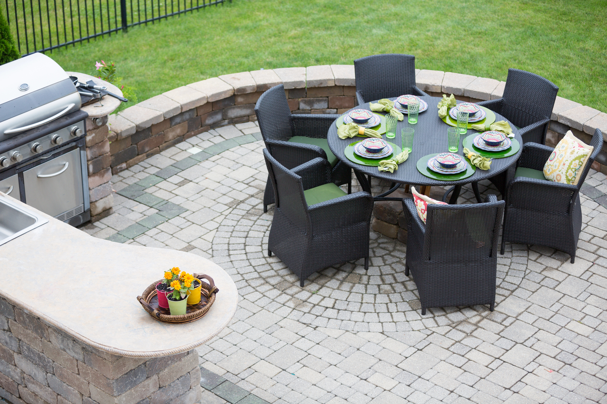 A round patio table set for six with green napkins sits on a stone patio near a grill and an outdoor counter with a small flowerpot and basket. Lawn and fence are in the background.