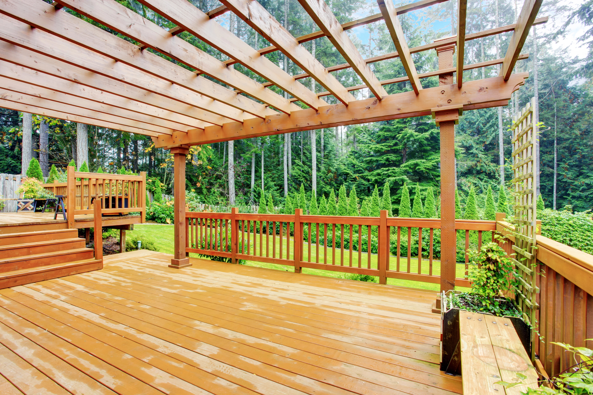Spacious wooden deck with a pergola, surrounded by a railing and lush green trees in the background.