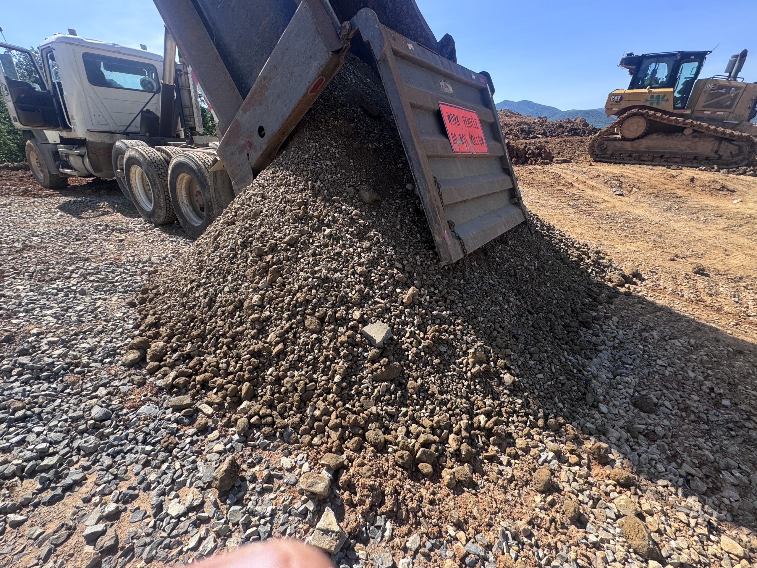 A dump truck unloads a pile of gravel onto a construction site, with a bulldozer in the background on a clear, sunny day.