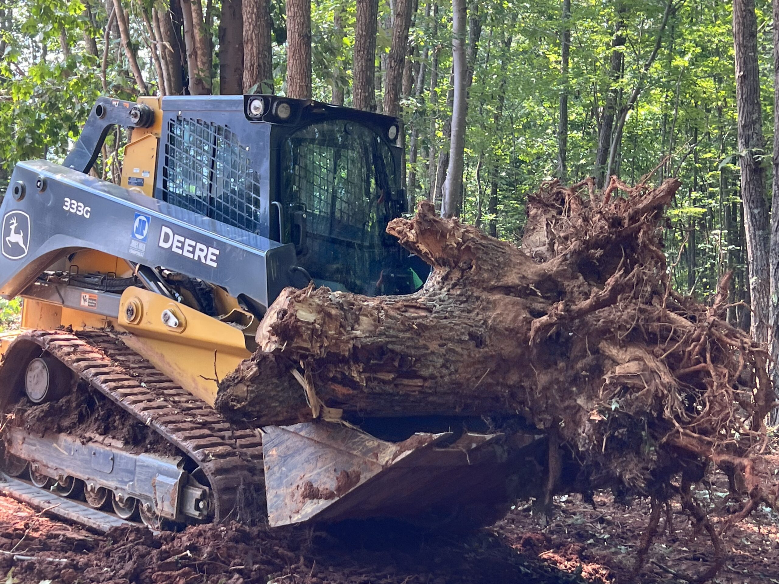 A John Deere skid steer loader lifts a large tree stump with roots exposed in a forest clearing.