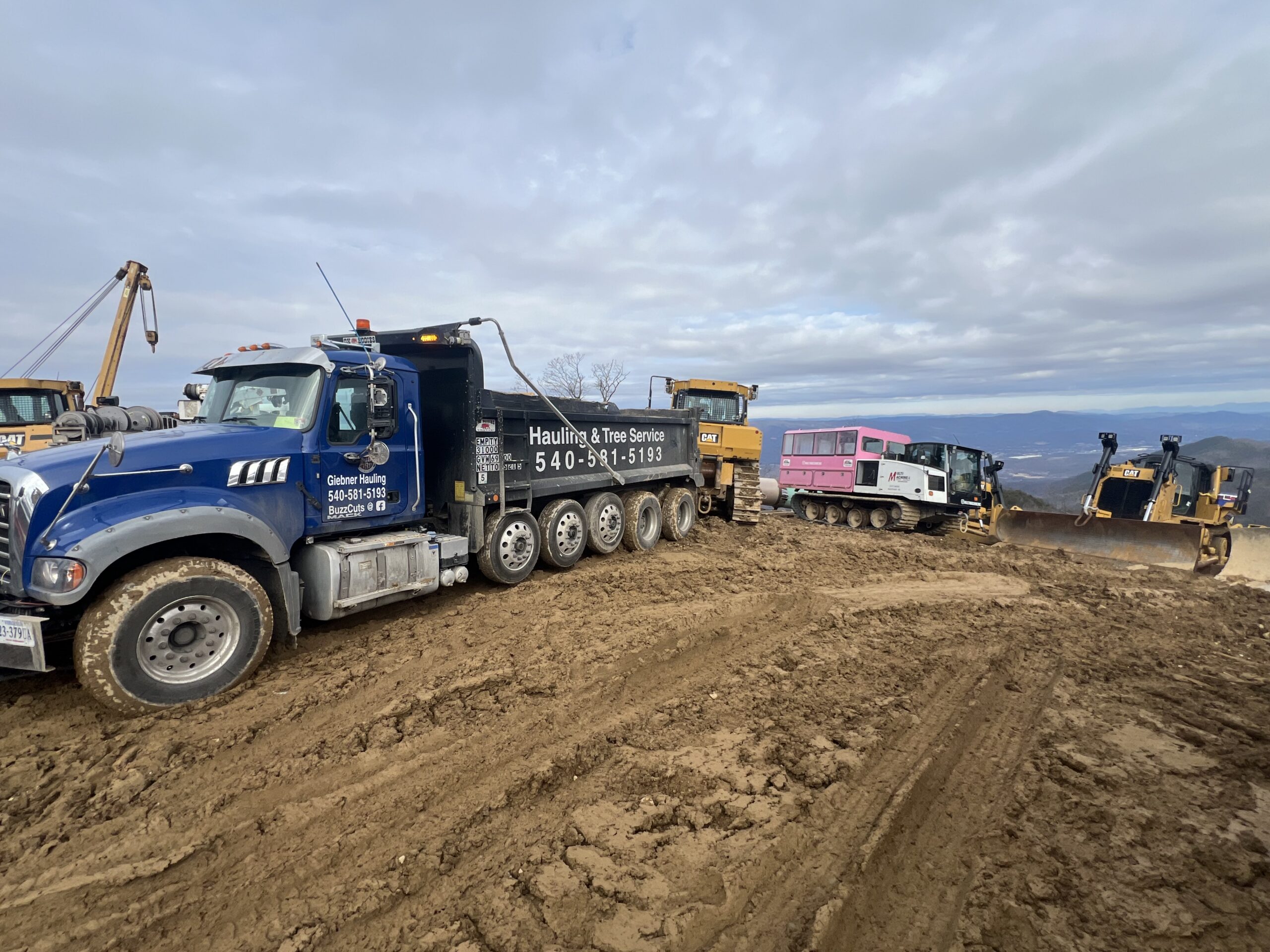 A large tow truck pulls a pink and white bus uphill on a muddy construction site, with bulldozers and machinery in the background under a cloudy sky.