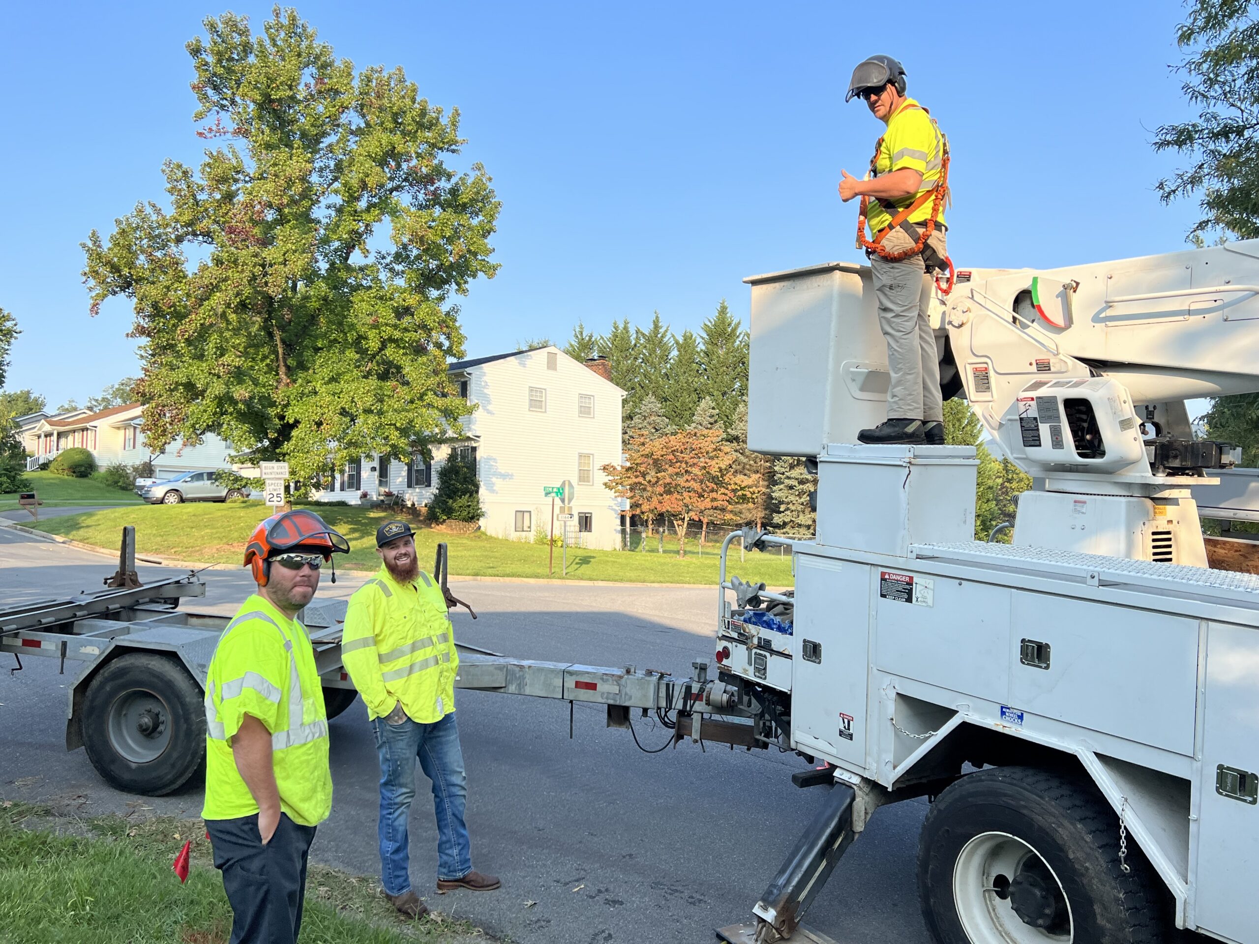 Three utility workers in safety gear stand near a utility truck with a bucket lift on a suburban street, with houses and trees in the background.
